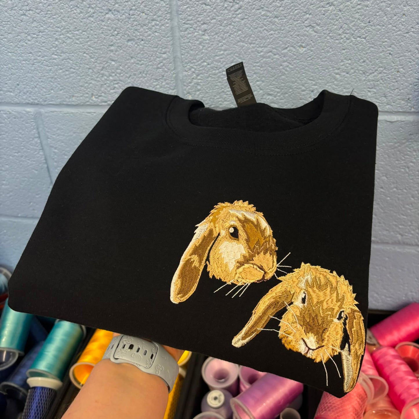 A black crewneck sweatshirt featuring two embroidered golden-brown rabbits on the front, held up by a person's hand in front of a gray brick wall and colorful spools of thread.