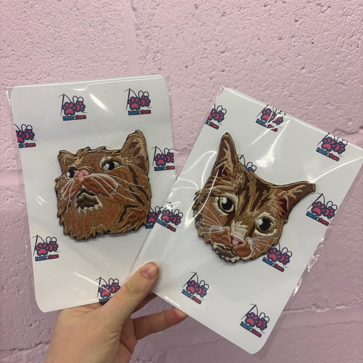 Two embroidered cat face patches on white backing cards with a repeating logo, held by a hand against a pink painted brick wall.