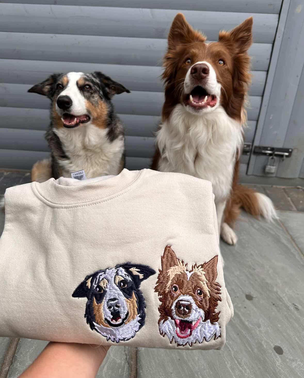 A folded cream sweatshirt held in a hand, featuring embroidered dog faces on the front, with two dogs sitting behind it against a gray metal door.