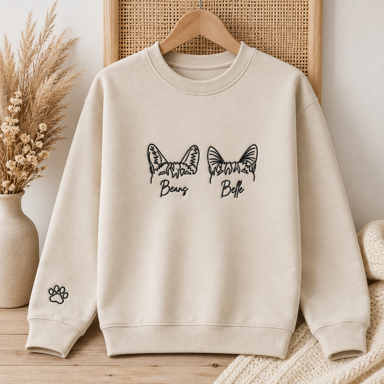 A cream-colored crewneck sweatshirt with black graphic prints of butterfly ears labeled 'Beaus' and 'Bells', displayed on a wooden hanger against a warm, neutral background with dried flowers and a woven cane panel.