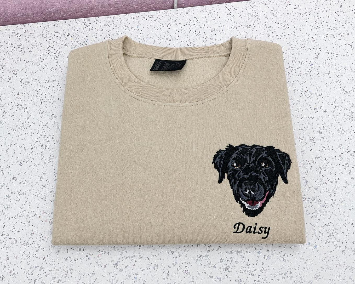 A beige crewneck sweatshirt folded flat on a speckled white countertop, featuring a black embroidered dog face and the name 'Daisy' printed below it.