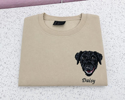 A beige crewneck sweatshirt folded flat on a speckled white countertop, featuring a black embroidered dog face and the name 'Daisy' printed below it.