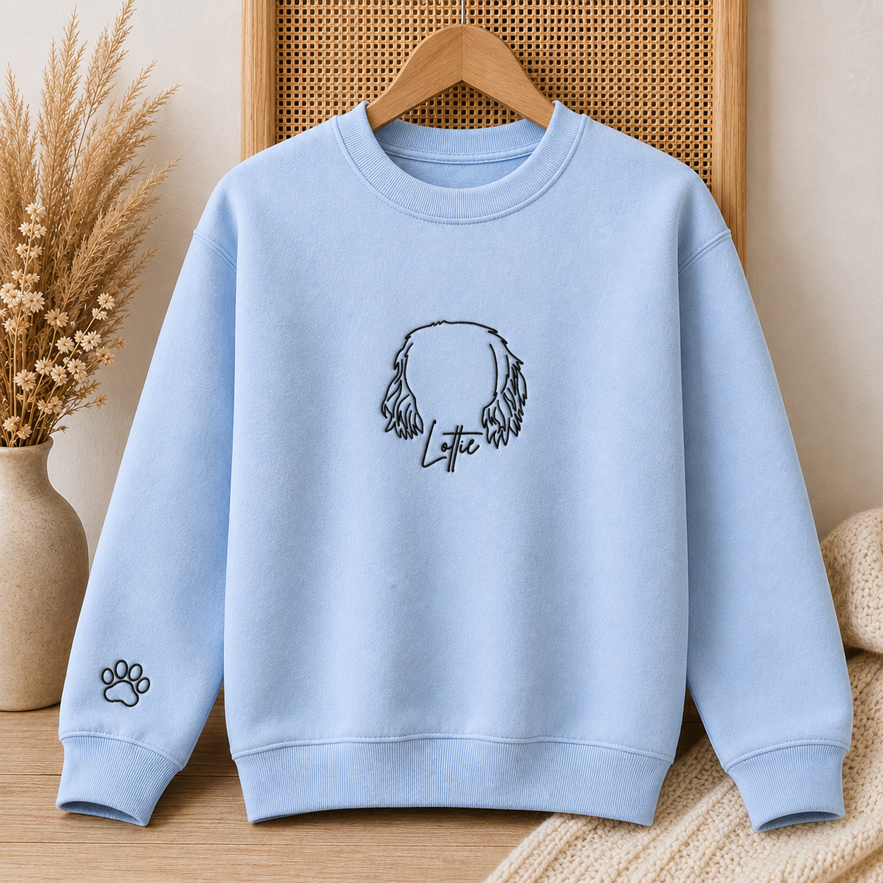 A light blue crewneck sweatshirt hanging on a wooden hanger, featuring a black line art graphic of a dog's head with the name 'Lytte' written below it, and a small black paw print on the left sleeve. The sweatshirt is displayed against a warm, neutral background with a woven panel and dried grass in a vase.