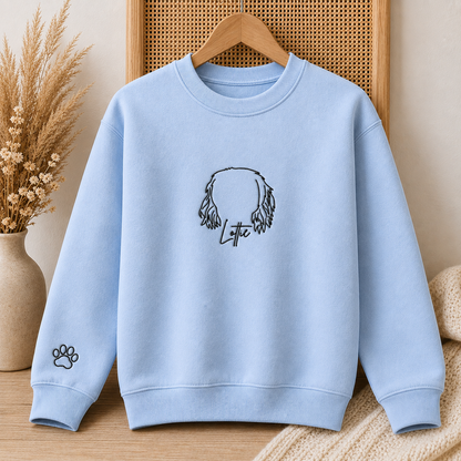 A light blue crewneck sweatshirt hanging on a wooden hanger, featuring a black line art graphic of a dog's head with the name 'Lytte' written below it, and a small black paw print on the left sleeve. The sweatshirt is displayed against a warm, neutral background with a woven panel and dried grass in a vase.