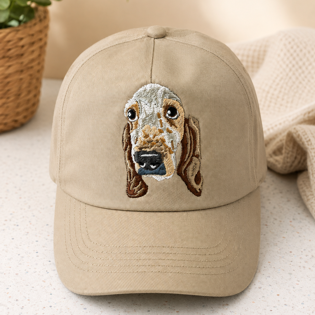 A beige baseball cap featuring an embroidered dog face on the front, styled on a light speckled surface with a potted plant and a textured throw blanket in the background.