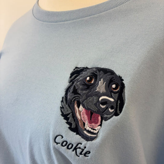A close-up of a light blue crewneck sweatshirt on a mannequin, featuring a detailed black and white embroidered dog head with its mouth open, and the name 'Cookie' embroidered below in black script.