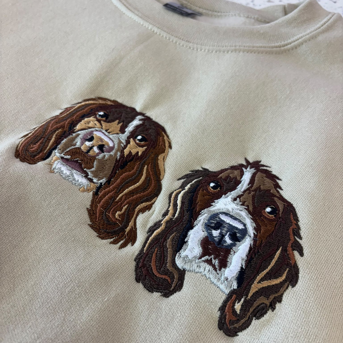Close-up of a cream-colored sweatshirt featuring two embroidered dog faces in brown, white, and black on the chest.