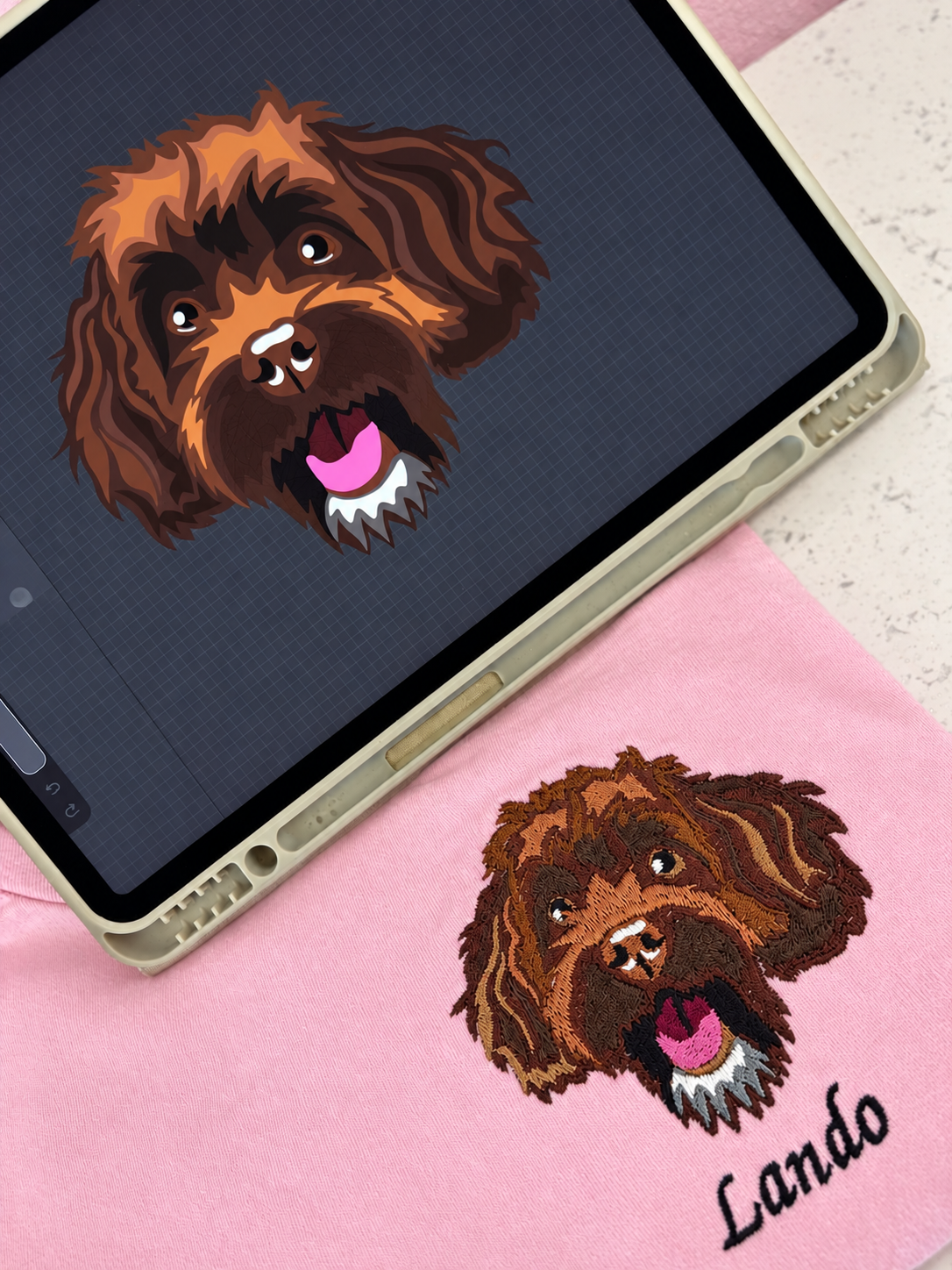 A pink embroidered pet portrait product shown on a light surface, with a tablet displaying the same dog-face design. The embroidery features a brown and tan dog head with pink tongue and white fur details, and the name 'Lando' embroidered below.