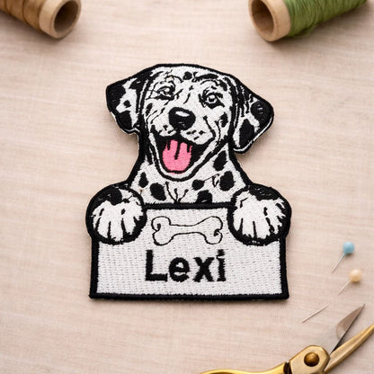 Embroidered patch of a dog holding a bone with 'Lexi' text, surrounded by sewing materials on a beige background.