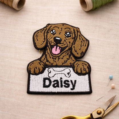 Embroidered patch of a dog with a name tag 'Daisy' on a fabric background with sewing tools.