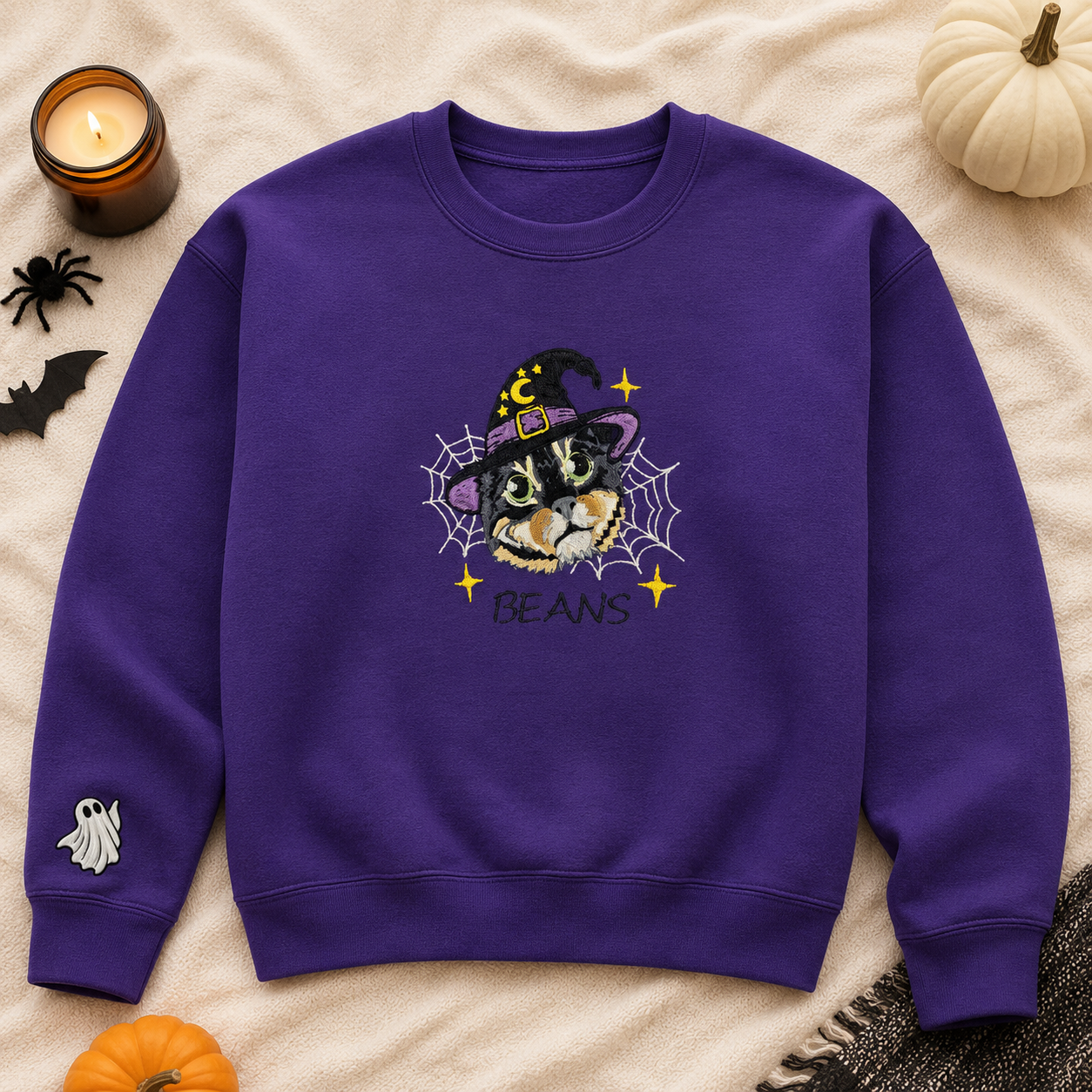 A purple crewneck sweatshirt laid flat on a beige fabric background with Halloween props. The sweatshirt features a graphic of a cat wearing a witch hat behind a spiderweb with the word 'BEANS' below, and a small white ghost motif on the left sleeve.