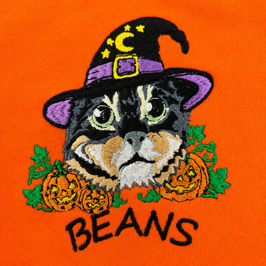 Close-up of an embroidered graphic on bright orange fabric featuring a black cat wearing a purple witch hat with yellow stars and a gold buckle, surrounded by orange pumpkins and green leaves, with the word "BEANS" embroidered below in black.