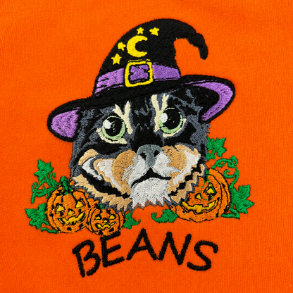 Close-up of an embroidered graphic on bright orange fabric featuring a black cat wearing a purple witch hat with yellow stars and a gold buckle, surrounded by orange pumpkins and green leaves, with the word "BEANS" embroidered below in black.