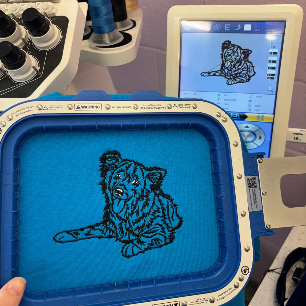 Embroidery machine with a blue embroidery frame featuring a tiger design on a screen displaying more designs.