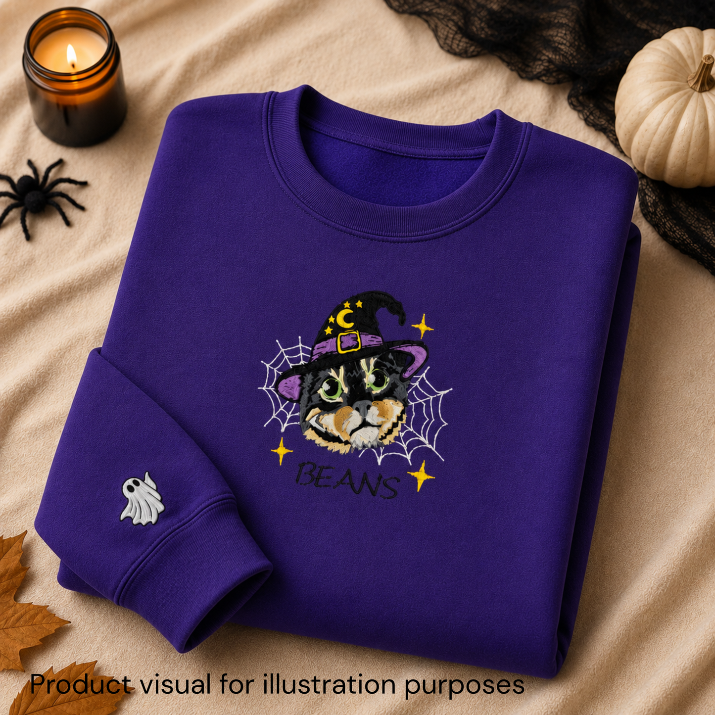 A folded purple sweatshirt with a Halloween-themed cat graphic on the front, styled on a beige fabric background with a candle, spider toy, black lace, pumpkin, and autumn leaf. The graphic shows a cat in a witch hat surrounded by a spiderweb with the text 'BEANS'.