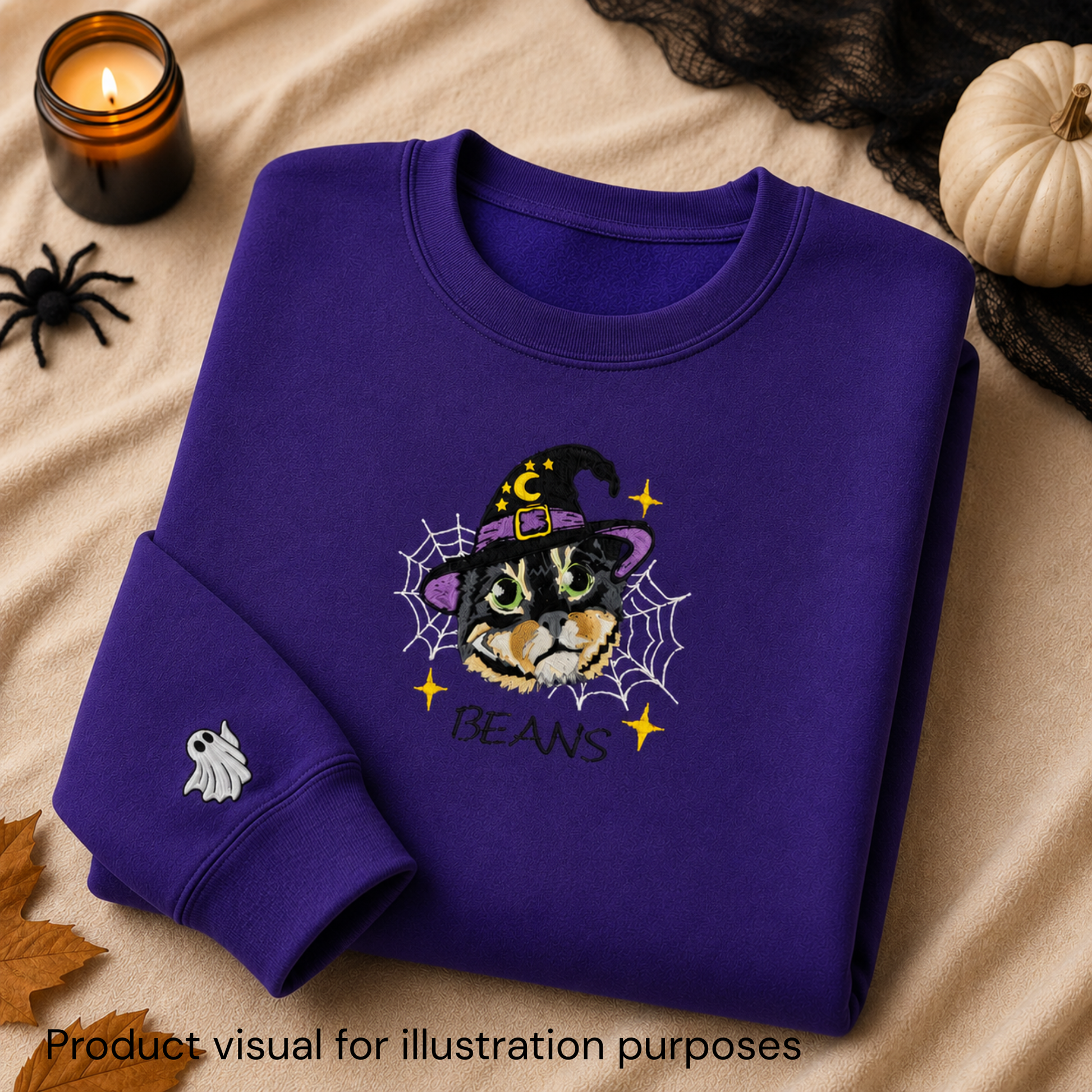 A folded purple sweatshirt with a Halloween-themed cat graphic on the front, styled on a beige fabric background with a candle, spider toy, black lace, pumpkin, and autumn leaf. The graphic shows a cat in a witch hat surrounded by a spiderweb with the text 'BEANS'.