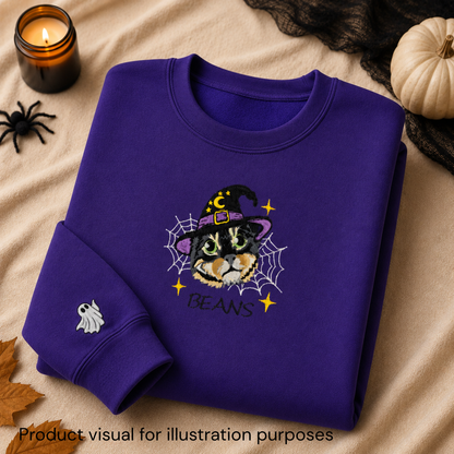A folded purple sweatshirt with a Halloween-themed cat graphic on the front, styled on a beige fabric background with a candle, spider toy, black lace, pumpkin, and autumn leaf. The graphic shows a cat in a witch hat surrounded by a spiderweb with the text 'BEANS'.