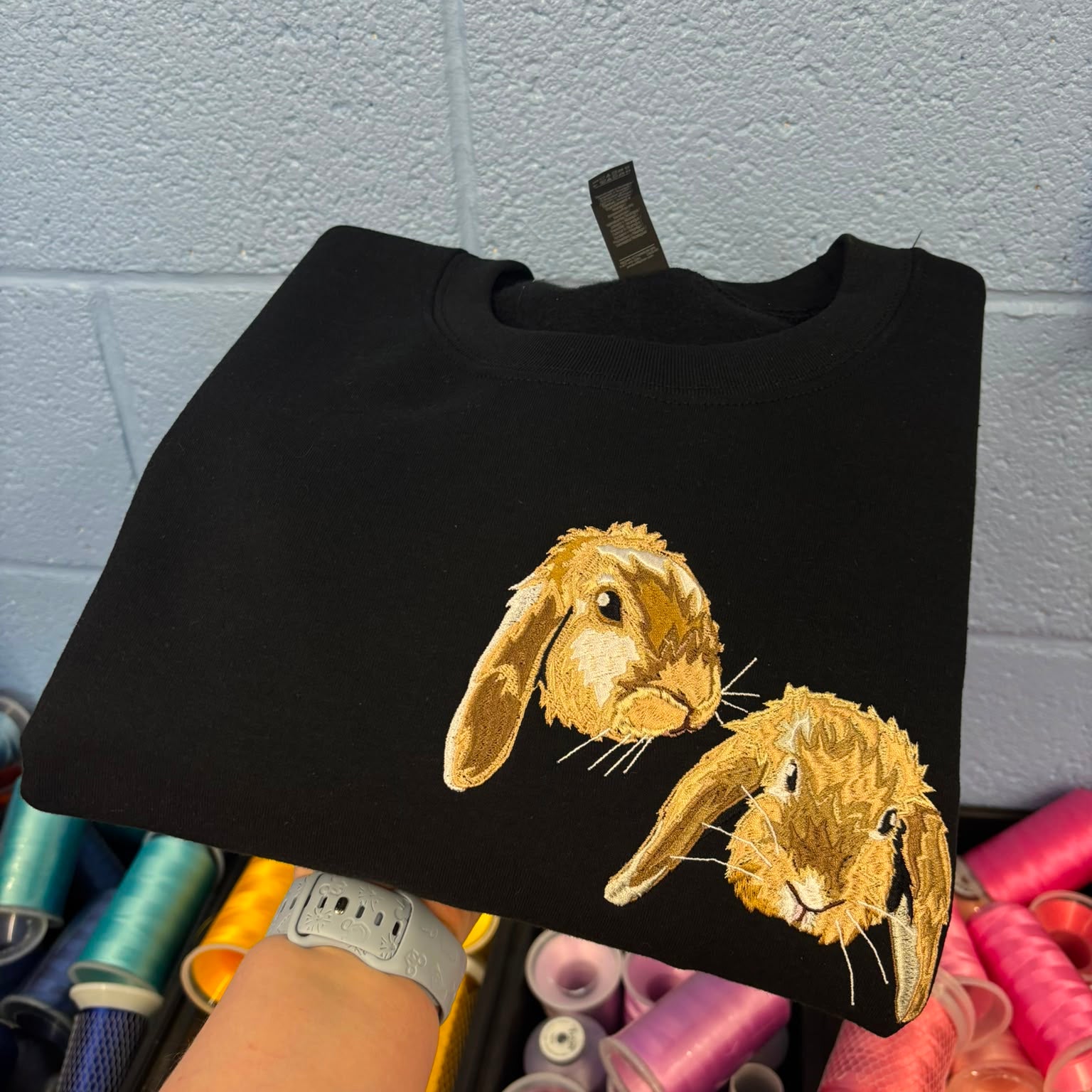 A black crewneck sweatshirt featuring two embroidered golden-brown rabbits on the front, held up by a person's hand in front of a gray brick wall and colorful spools of thread.