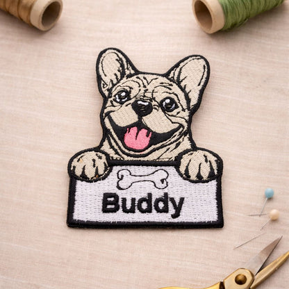Embroidered patch of a dog with 'Buddy' text on a fabric background with sewing tools.